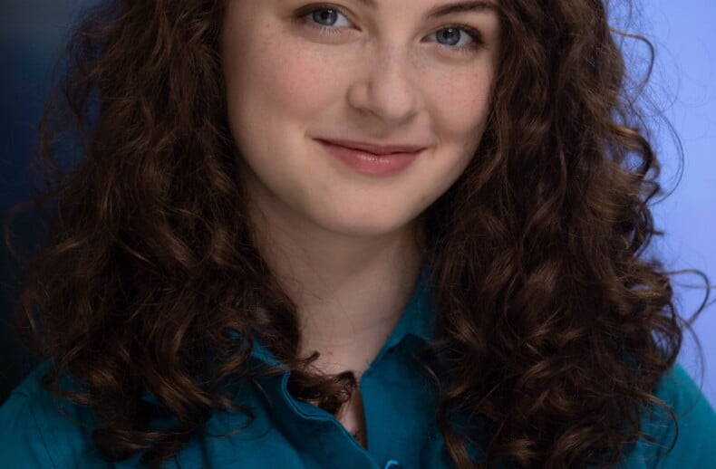 A person with long, curly brown hair smiles gently at the camera. They wear a teal shirt against a blurred blue background, reflecting the welcoming and inclusive spirit of diversity programs while presenting a professional and friendly appearance.