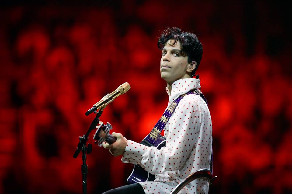A musician strums his guitar on stage, clad in a white polka-dotted shirt, with a golden microphone before him. The vibrant red backdrop enhances the dramatic atmosphere, mirroring the emotional depth found in social emotional learning curriculum.