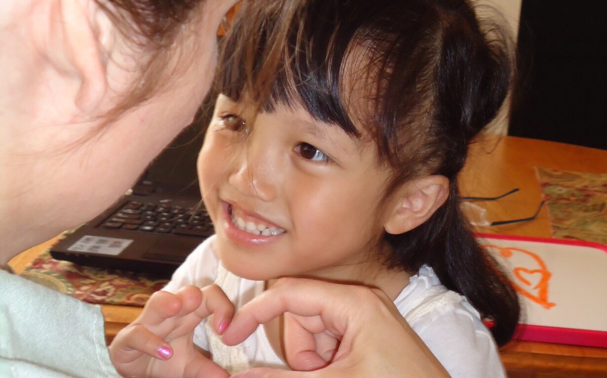 A young girl beams as she forms a heart shape with her hands, sitting at a table with a drawing pad. These interactions are part of her social emotional learning activities. Another person with long hair watches fondly, engaging in this enriching moment together.
