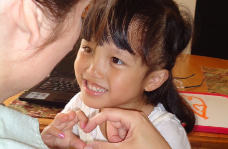 A young girl beams as she forms a heart shape with her hands, sitting at a table with a drawing pad. These interactions are part of her social emotional learning activities. Another person with long hair watches fondly, engaging in this enriching moment together.