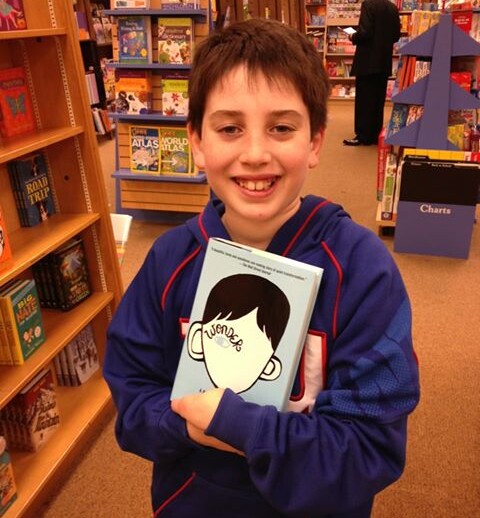 In a cozy bookstore, a young boy beams with joy, holding the blue-covered book "Wonder," as he explores shelves brimming with stories that inspire SEL activities.
