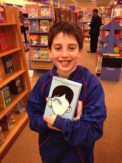 In a cozy bookstore, a young boy beams with joy, holding the blue-covered book "Wonder," as he explores shelves brimming with stories that inspire SEL activities.