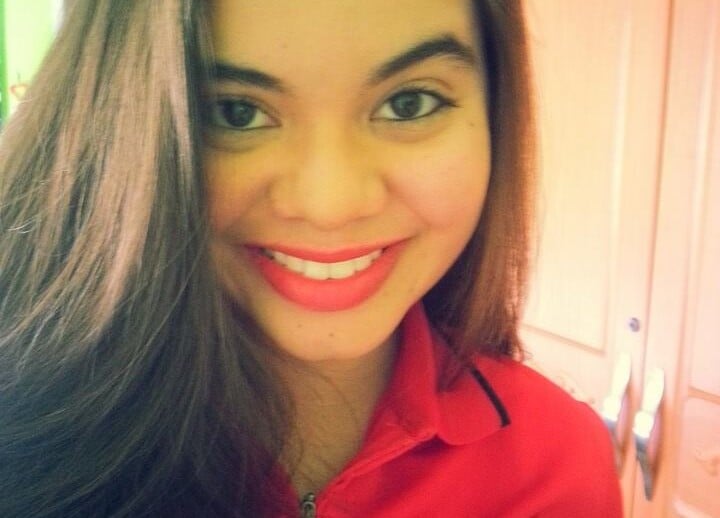 A woman with long brown hair smiles at the camera, radiating warmth like the engaging activities found in an SEL curriculum. She is wearing a red top against a backdrop of a green wall and wooden wardrobe, creating an inviting atmosphere for learning and growth.