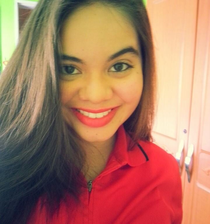 A woman with long brown hair smiles at the camera, radiating warmth like the engaging activities found in an SEL curriculum. She is wearing a red top against a backdrop of a green wall and wooden wardrobe, creating an inviting atmosphere for learning and growth.