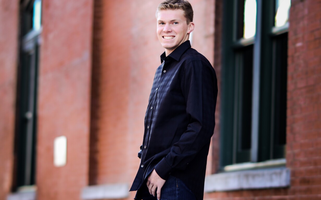 A person in a black shirt and blue jeans, showcasing the spirit of diversity programs, smiles while standing in front of a brick wall with large windows.