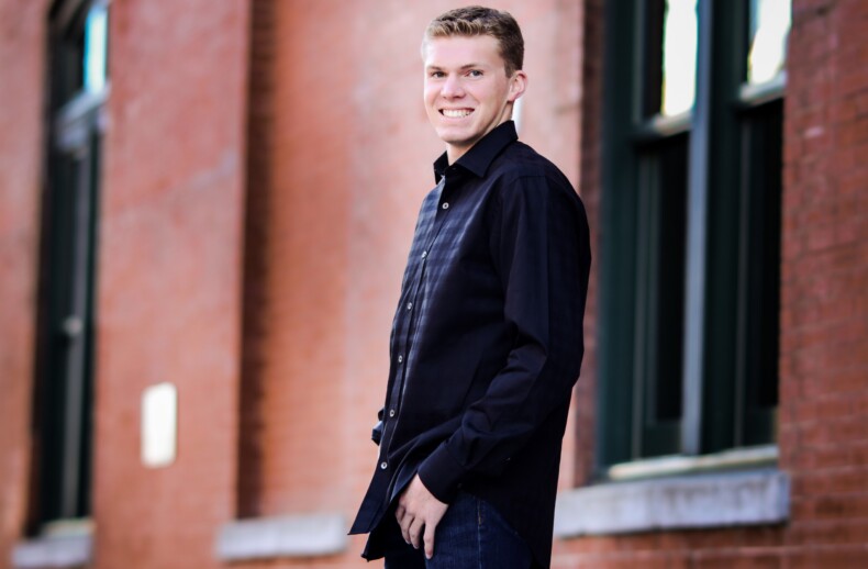 A person in a black shirt and blue jeans, showcasing the spirit of diversity programs, smiles while standing in front of a brick wall with large windows.
