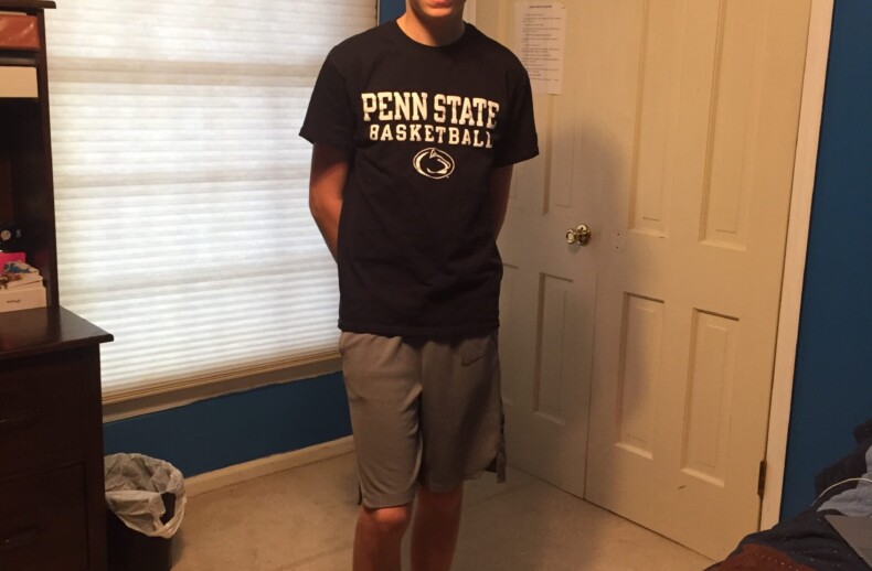 Teen in a cap, Penn State basketball shirt, and shorts stands in a room with blue walls. A small basketball hoop hangs on the door, reminiscent of a space fostering teamwork found in SEL curriculum. The bed is partially visible on the right with a colorful quilt.