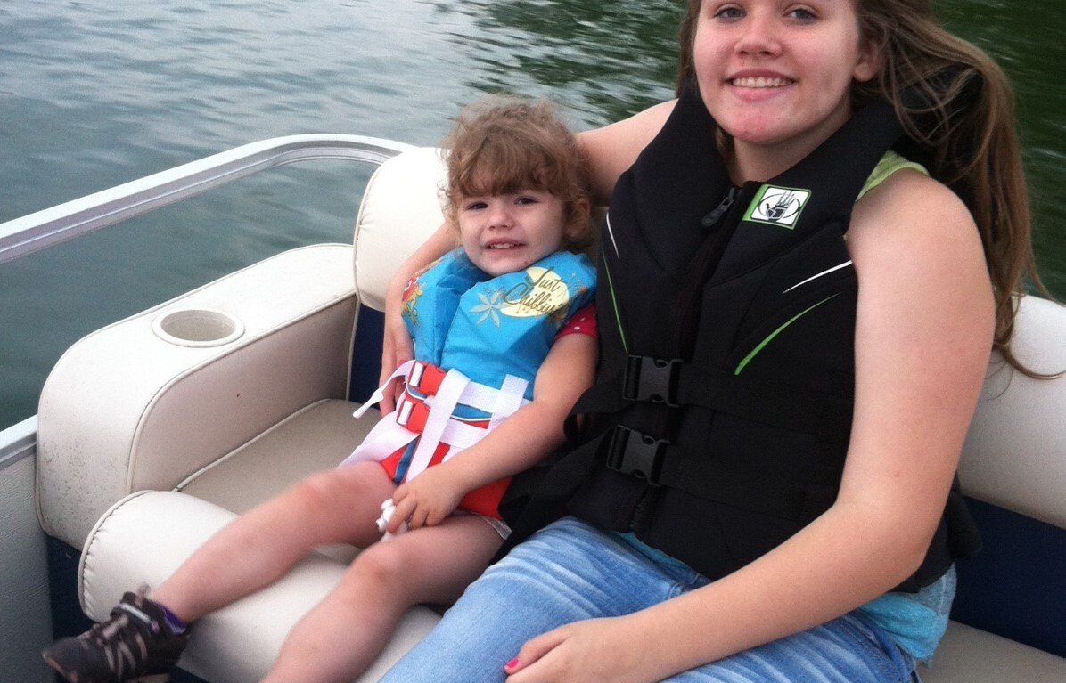A young woman in a life jacket sits on a boat seat, smiling at the camera. Beside her, a girl in a blue shirt and colorful shorts reclines, also smiling. This serene moment on calm waters amidst lush greenery embodies the spirit of SEL activities—a perfect setting for connection and understanding.