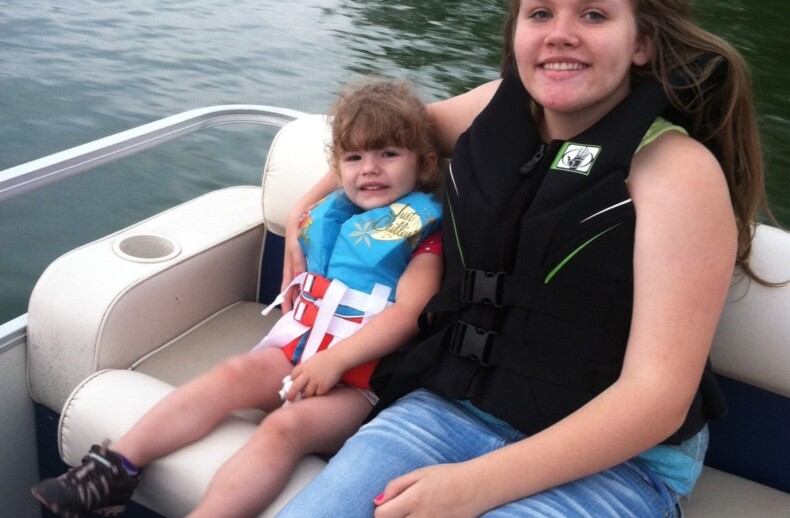 A young woman in a life jacket sits on a boat seat, smiling at the camera. Beside her, a girl in a blue shirt and colorful shorts reclines, also smiling. This serene moment on calm waters amidst lush greenery embodies the spirit of SEL activities—a perfect setting for connection and understanding.