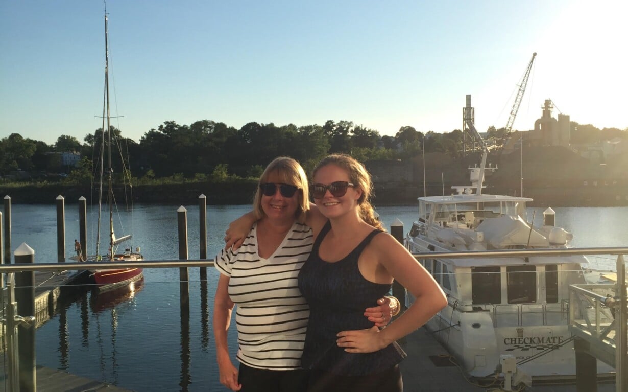 Two people stand smiling on a waterfront deck under a clear blue sky. A dock and boats are visible in the background, with trees lining the distant shore. Both are wearing sunglasses, enjoying the sunny day, much like one spent in engaging SEL activities that foster connection and joy.