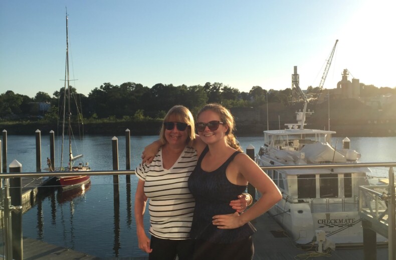 Two people stand smiling on a waterfront deck under a clear blue sky. A dock and boats are visible in the background, with trees lining the distant shore. Both are wearing sunglasses, enjoying the sunny day, much like one spent in engaging SEL activities that foster connection and joy.