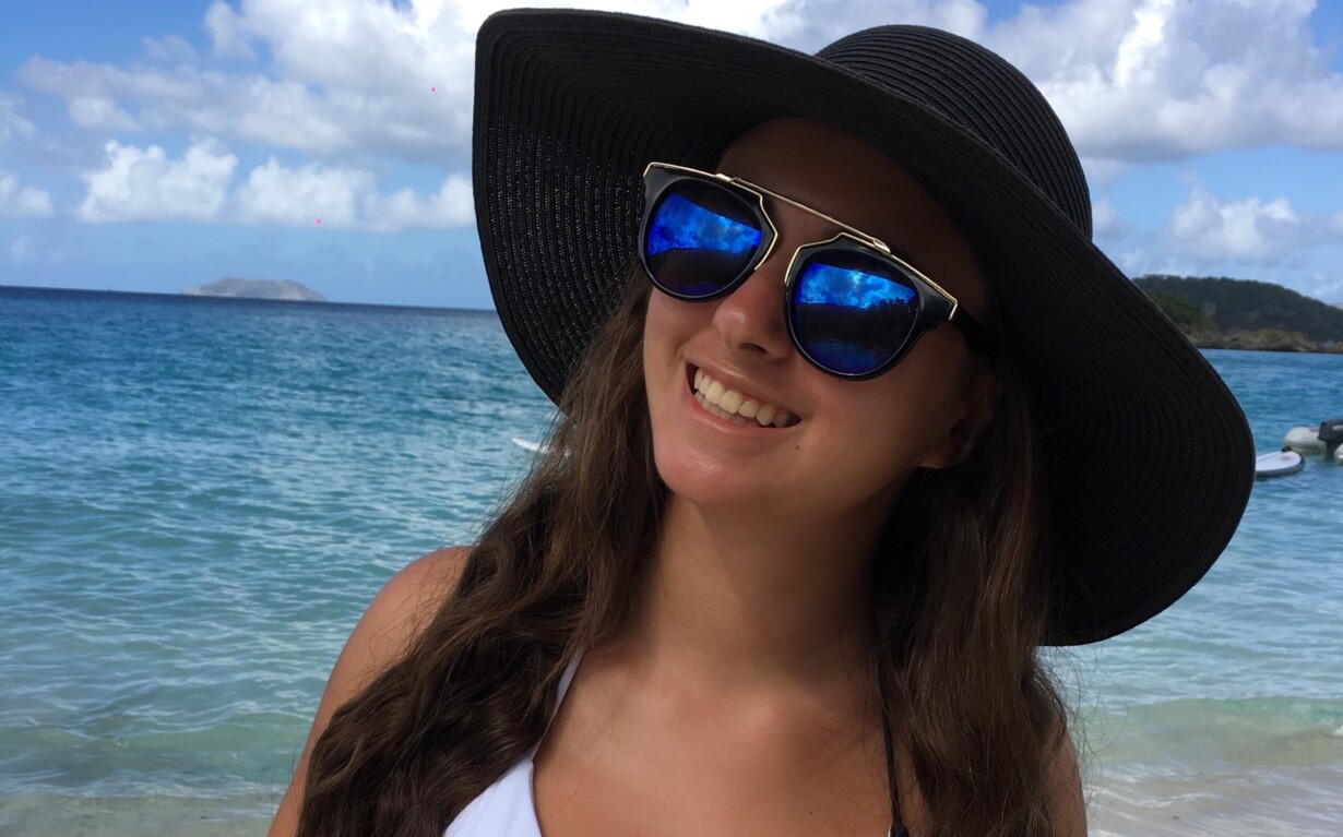 A woman in a black sunhat and sunglasses smiles on the beach, embodying the carefree essence of a well-planned SEL curriculum. She wears a black and white swimsuit, with serene blue ocean waves and a cloudy sky providing the ideal backdrop for introspective get to know you activities.
