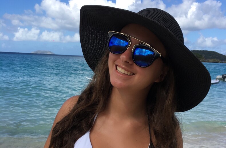 A woman in a black sunhat and sunglasses smiles on the beach, embodying the carefree essence of a well-planned SEL curriculum. She wears a black and white swimsuit, with serene blue ocean waves and a cloudy sky providing the ideal backdrop for introspective get to know you activities.