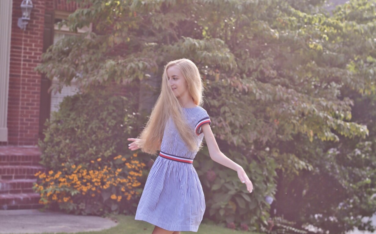 A woman with long blonde hair spins joyfully on a grassy lawn in front of a brick house. She is barefoot, wearing a light blue dress. Sunlight filters through nearby trees, casting a serene glow—a scene perfect for inspiring moments within an SEL curriculum.