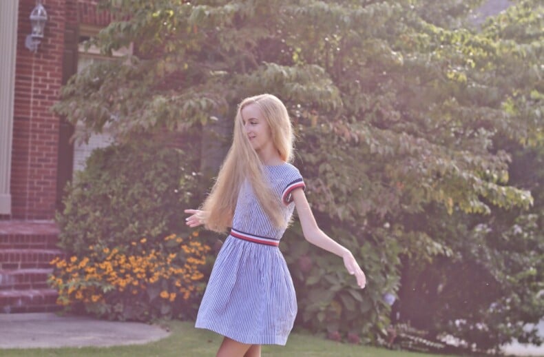 A woman with long blonde hair spins joyfully on a grassy lawn in front of a brick house. She is barefoot, wearing a light blue dress. Sunlight filters through nearby trees, casting a serene glow—a scene perfect for inspiring moments within an SEL curriculum.