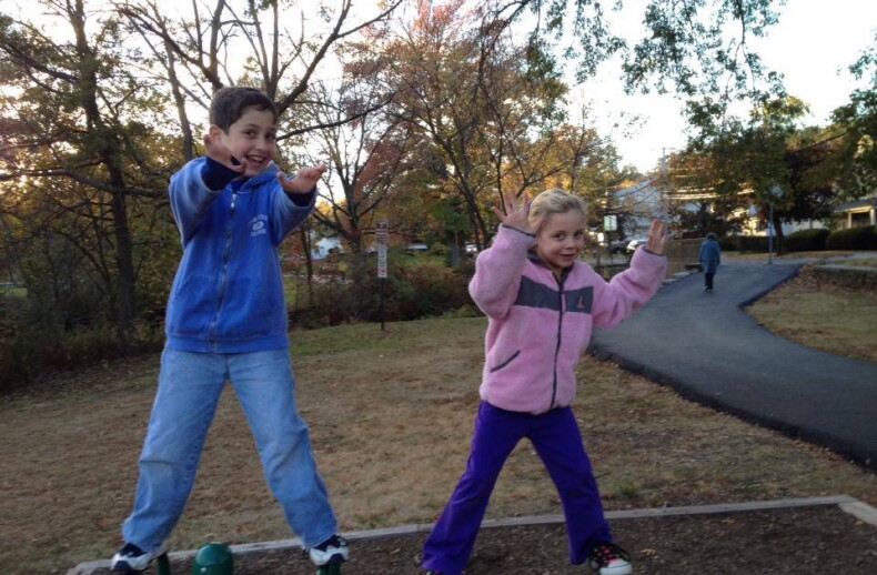 Two children stand on green bars in a park, playfully posing with arms outstretched as part of an engaging SEL curriculum. They wear warm clothes, one in a blue jacket and the other in pink. Autumn trees and a pathway paint a picturesque backdrop to their social emotional learning activities.