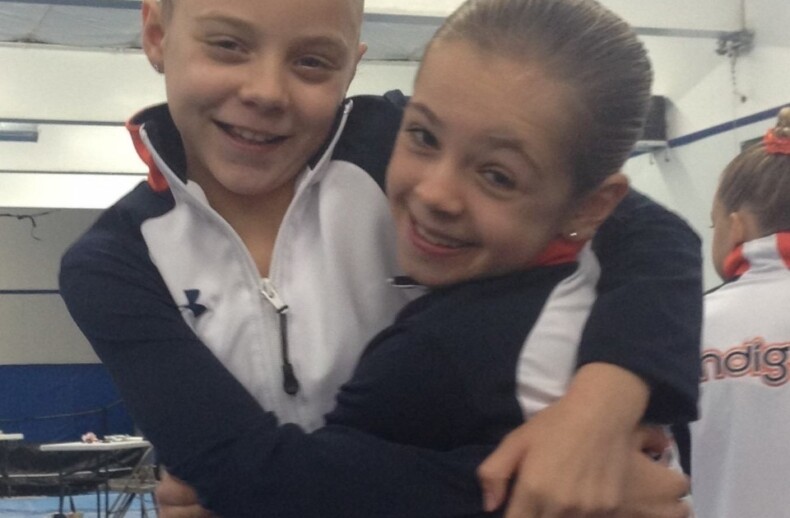 Two young gymnasts wearing matching black and white tracksuits with orange accents hug each other and smile, embodying the spirit of teamwork fostered through social emotional learning activities. They stand in a gymnasium with blue mats and equipment in the background.