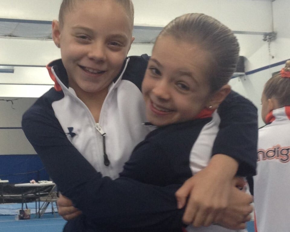 Two young gymnasts wearing matching black and white tracksuits with orange accents hug each other and smile, embodying the spirit of teamwork fostered through social emotional learning activities. They stand in a gymnasium with blue mats and equipment in the background.