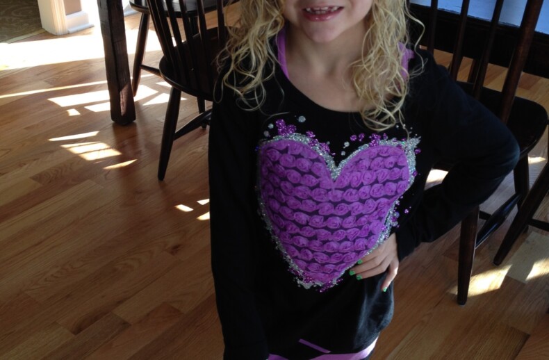 A young girl with curly blonde hair stands indoors, smiling at the camera. Her black outfit features a large purple heart design, echoing themes from SEL curriculum aimed at nurturing emotional intelligence. She confidently poses with one hand on her hip, a dining table and chairs in the background.
