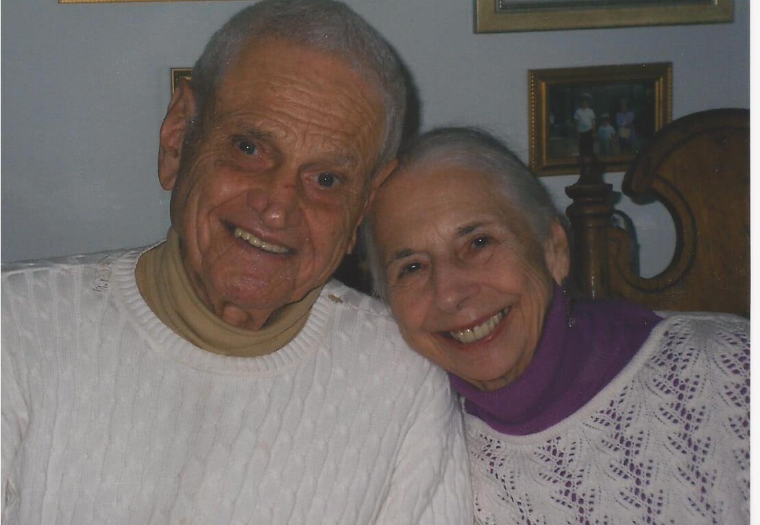 An elderly couple, both with white hair, smile warmly at the camera, embodying the essence of social emotional learning activities. They are wearing matching white knit tops. The background shows framed pictures on the wall.