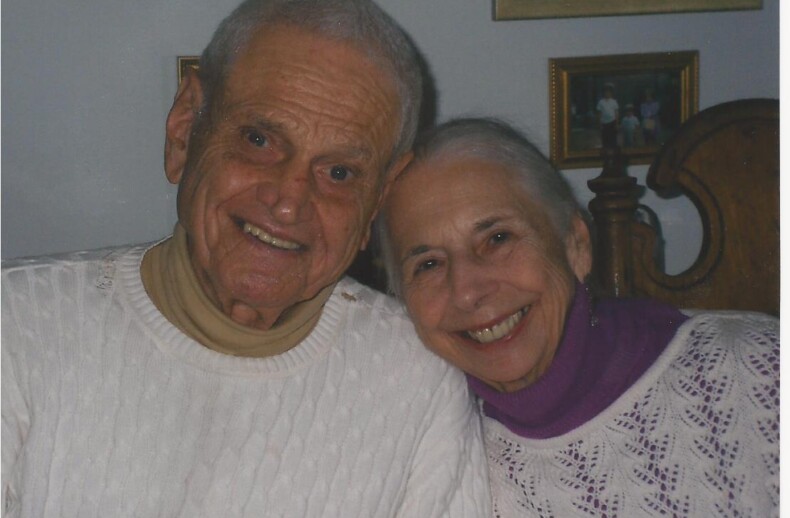 An elderly couple, both with white hair, smile warmly at the camera, embodying the essence of social emotional learning activities. They are wearing matching white knit tops. The background shows framed pictures on the wall.