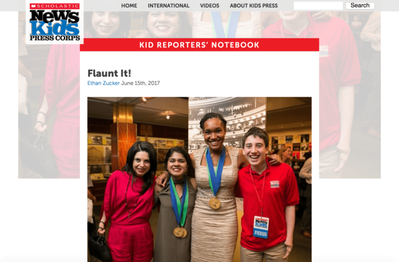 Four people stand smiling together in front of a display. One wears a formal dress and two medals, another has a press pass, and two are in evening attire. The magazine-themed banner, "Scholastic News Kids Press Corps," highlights the event's focus on diversity programs.