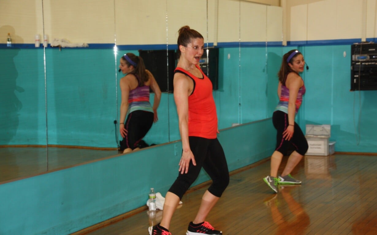 A woman in a red tank top and black leggings energetically performs a dance exercise in a mirrored studio, integrating social emotional learning activities. She wears a headset, colorful sneakers, and looks enthusiastic. Another person is visible in the mirror reflection behind her.