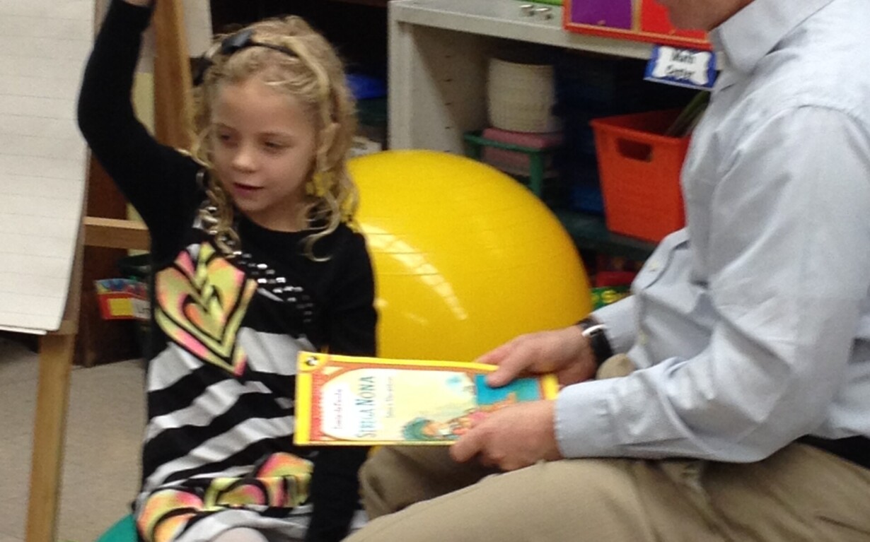 A man sits on a chair holding a book, engaging in social emotional learning activities, while a young girl on an exercise ball raises her hand. They are in a classroom filled with educational materials, fostering an environment enriched by a thoughtful SEL curriculum.