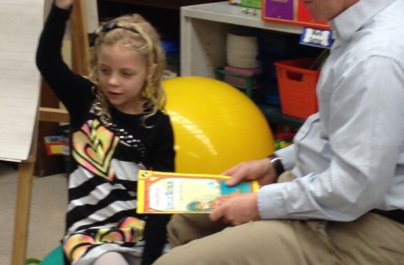 A man sits on a chair holding a book, engaging in social emotional learning activities, while a young girl on an exercise ball raises her hand. They are in a classroom filled with educational materials, fostering an environment enriched by a thoughtful SEL curriculum.