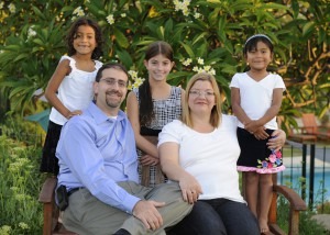 A family of five poses outdoors in a garden, embodying the spirit of social emotional learning activities. Two adults sit on a bench, with a man in a blue shirt and a woman in white. Three young girls, smiling brightly, stand behind them amidst lush greenery and vibrant flowers.