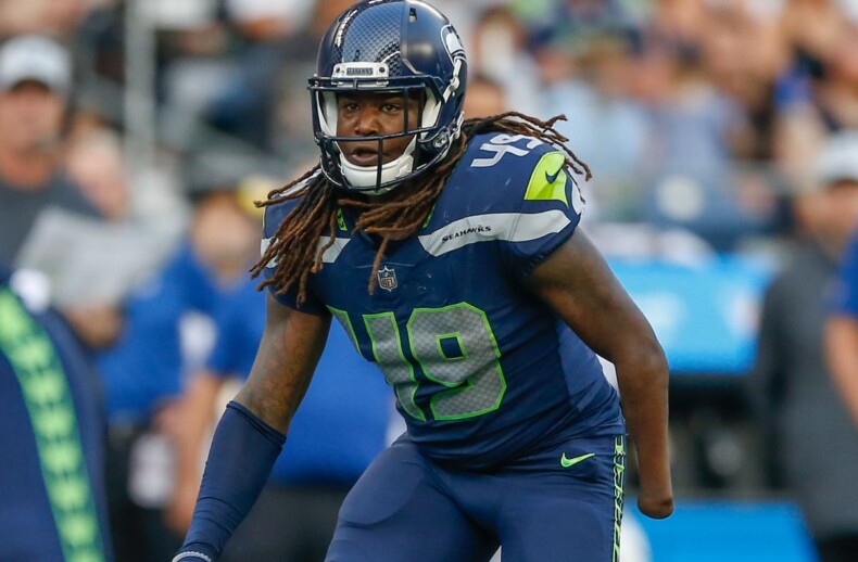 Football player in a Seattle Seahawks uniform on the field, with the number 49 on his jersey. Wearing a helmet, he appears focused during the game as diversity programs engage fans in the stands filled with spectators cheering in the background.