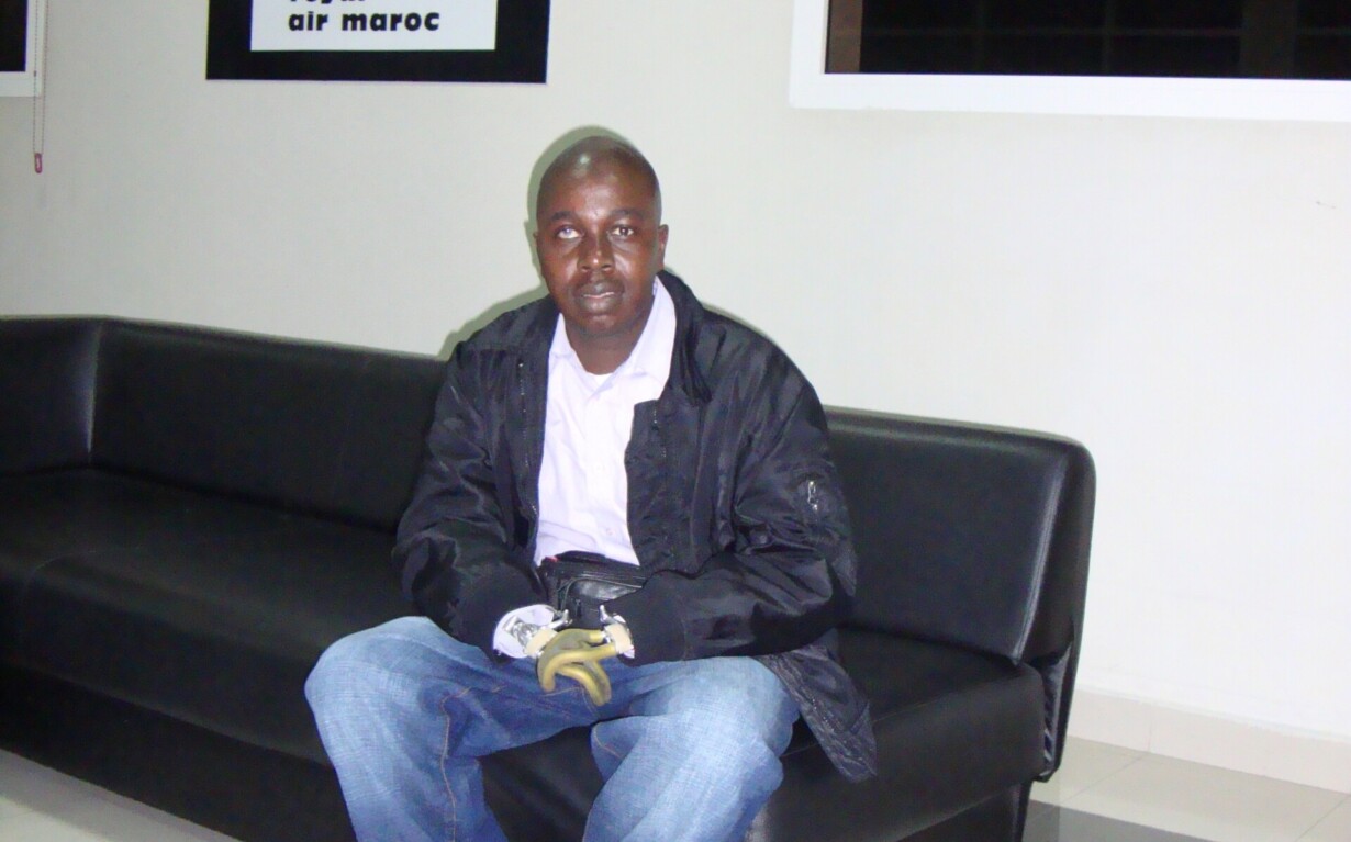 A man wearing a black jacket and jeans sits on a black sofa in a modern room, embodying the calm focus often nurtured by SEL activities. Behind him is a black and white "Royal Air Maroc" poster. The floor is tiled, and large windows bring natural light into this serene setting.