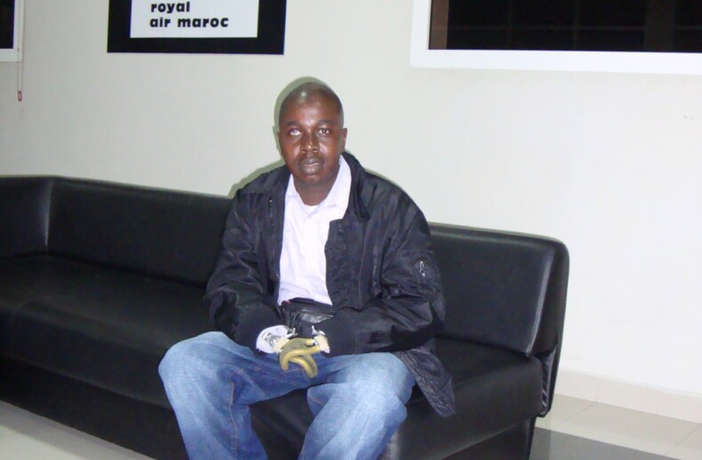 A man wearing a black jacket and jeans sits on a black sofa in a modern room, embodying the calm focus often nurtured by SEL activities. Behind him is a black and white "Royal Air Maroc" poster. The floor is tiled, and large windows bring natural light into this serene setting.