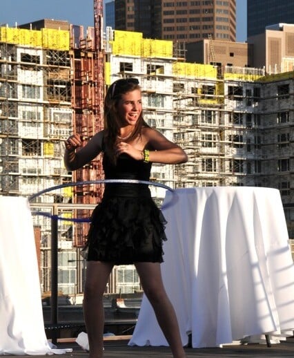 A young woman in a black dress hula hoops on a rooftop, seamlessly blending into the cityscape with unfinished buildings. She stands on a sunlit wooden deck, flanked by white tables covered with cloths—a perfect setting for embracing diversity programs in creative spaces.