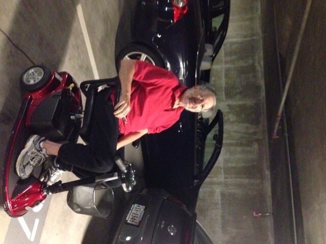 A man in a red shirt sits on a motorized mobility scooter in a parking garage, beside parked cars. The concrete walls echo quiet solitude, reminiscent of moments between social emotional learning activities, where one reflects amidst life's hustle. A car with a visible license plate stands nearby.