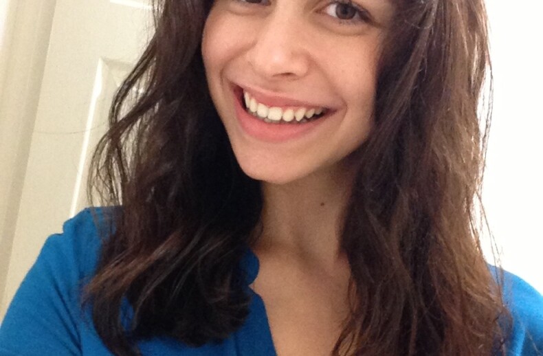 A person with long dark hair is smiling at the camera, embodying the joy often encouraged by social emotional learning activities. They are wearing a blue shirt and standing in front of a white door, radiating positivity and openness.