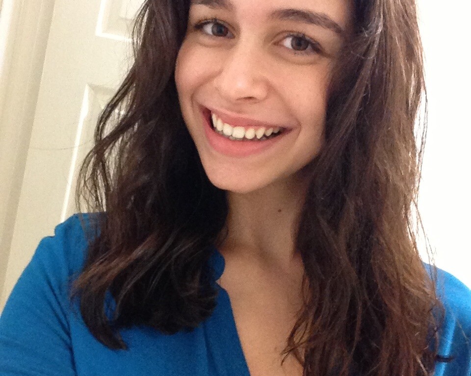 A person with long dark hair is smiling at the camera, embodying the joy often encouraged by social emotional learning activities. They are wearing a blue shirt and standing in front of a white door, radiating positivity and openness.