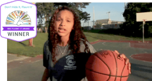 A person with long curly hair stands on a basketball court, holding a basketball. They're wearing a gray shirt, embodying the essence of social emotional learning activities. A badge on the image reads, "Don’t Hide It, Flaunt It - NBC Flaunt It Award WINNER." Trees and a basketball hoop create a vibrant backdrop.