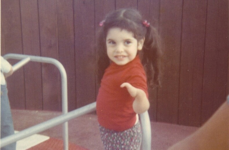 A young child with long dark hair in pigtails stands on a red playground merry-go-round, smiling at the camera. Wearing a red shirt and patterned pants, the child's joy reflects the essence of social emotional learning activities. A wooden fence serves as the background.