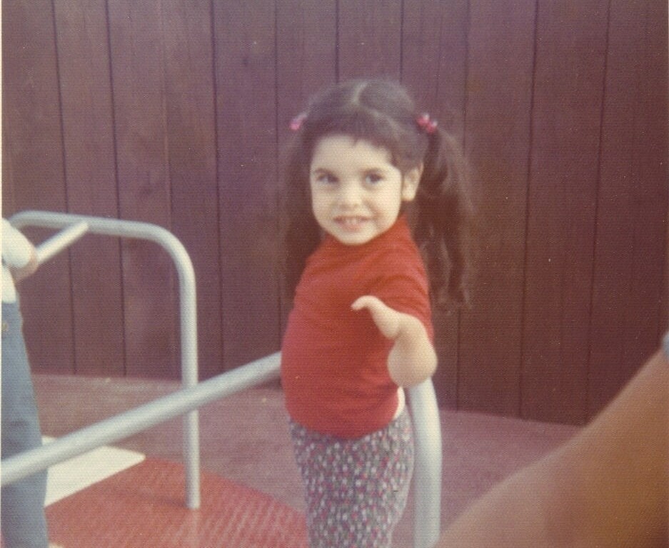 A young child with long dark hair in pigtails stands on a red playground merry-go-round, smiling at the camera. Wearing a red shirt and patterned pants, the child's joy reflects the essence of social emotional learning activities. A wooden fence serves as the background.