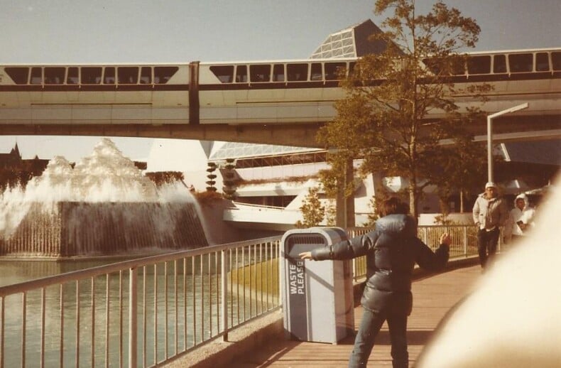 A person in a winter jacket stands animatedly on a walkway near a railing, with a monorail passing above. Embracing the diversity programs atmosphere, the scene reveals futuristic structures and a water feature in the background, while others stroll nearby.