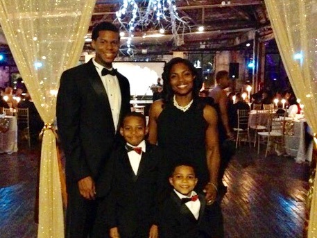 A family dressed in formal attire poses in an elegant venue, embodying the principles of diversity programs. Two adults and two children, all wearing black suits with red bow ties, stand under a canopy of lights and decorative curtains. A festive, warmly lit atmosphere surrounds them.