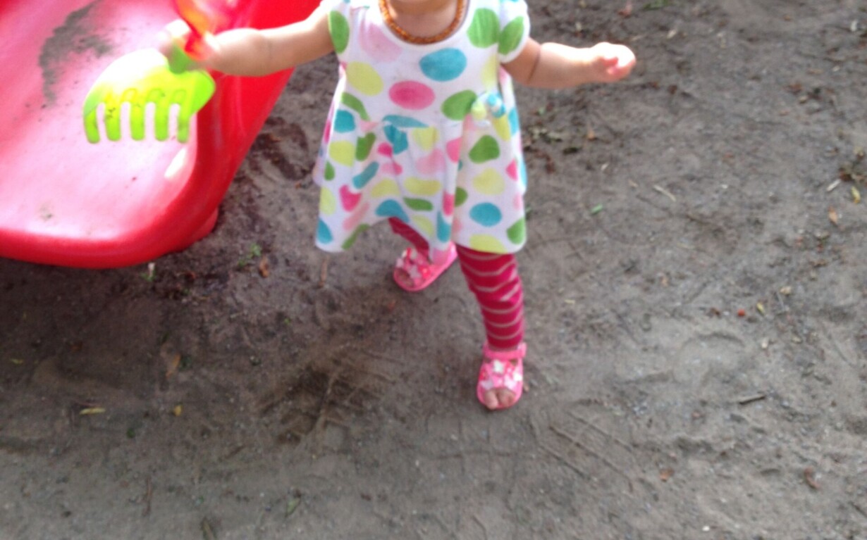 A toddler in a colorful polka dot dress and pink-striped leggings stands on sandy ground near a red slide, clutching a green toy rake—a playful scene that echoes the joy experienced during social emotional learning curriculum activities.