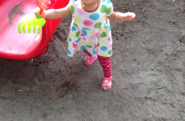 A toddler in a colorful polka dot dress and pink-striped leggings stands on sandy ground near a red slide, clutching a green toy rake—a playful scene that echoes the joy experienced during social emotional learning curriculum activities.