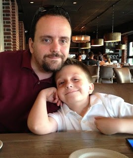 A man and a young boy are sitting at a restaurant table, engaging in meaningful talk akin to social emotional learning activities. The man has his arm around the boy, who is smiling and leaning against him. The background shows other diners and warm lighting.