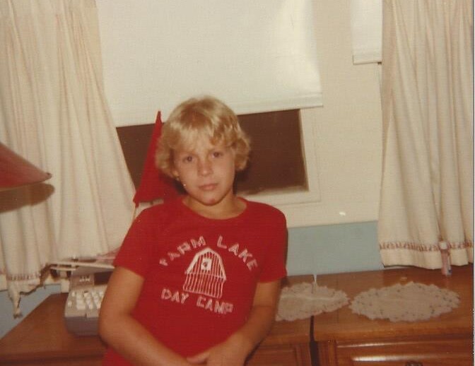 A young child with blonde hair, wearing a red "Farm Lake Day Camp" T-shirt, stands in a room with light-colored curtains and lace doilies on a dresser. Nearby, a red lamp and rotary phone hint at nostalgic charm within the space that encourages SEL activities for nurturing emotional growth.