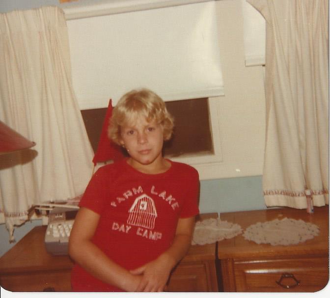 A young child with blonde hair, wearing a red "Farm Lake Day Camp" T-shirt, stands in a room with light-colored curtains and lace doilies on a dresser. Nearby, a red lamp and rotary phone hint at nostalgic charm within the space that encourages SEL activities for nurturing emotional growth.