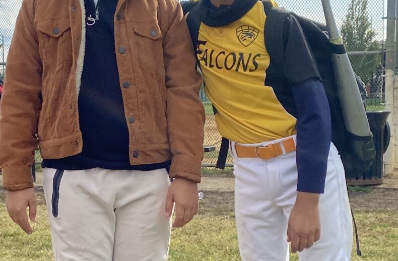 Two boys stand on a grassy field, embodying the spirit of social emotional learning activities. One wears a brown jacket and sunglasses, while the other sports a yellow and white "Falcons" baseball uniform with a bat in hand. Smiles shine under the cloudy sky, framed by a chain-link fence.
