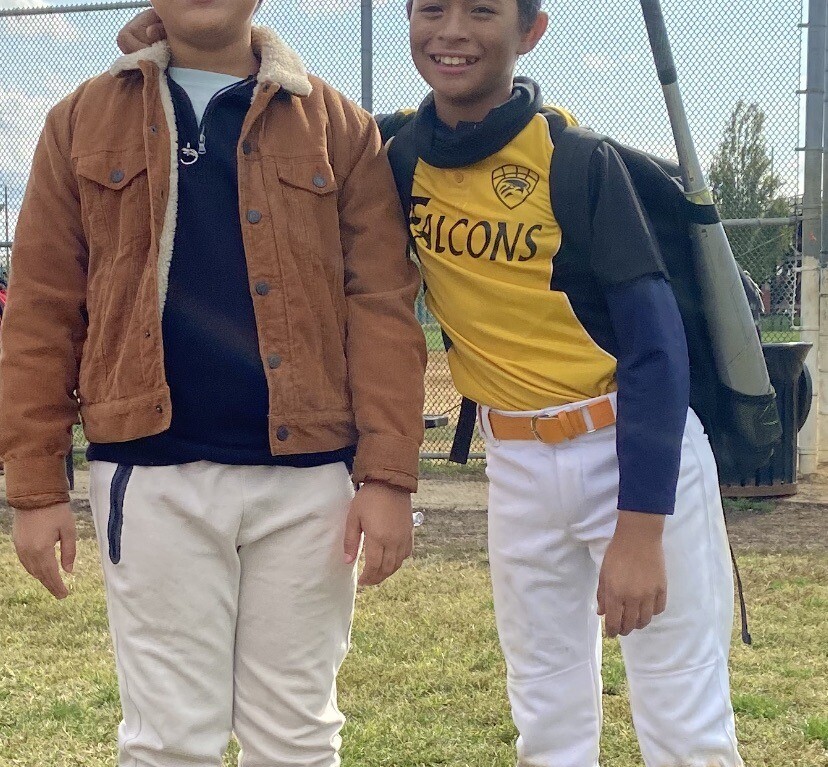 Two boys stand on a grassy field, embodying the spirit of social emotional learning activities. One wears a brown jacket and sunglasses, while the other sports a yellow and white "Falcons" baseball uniform with a bat in hand. Smiles shine under the cloudy sky, framed by a chain-link fence.