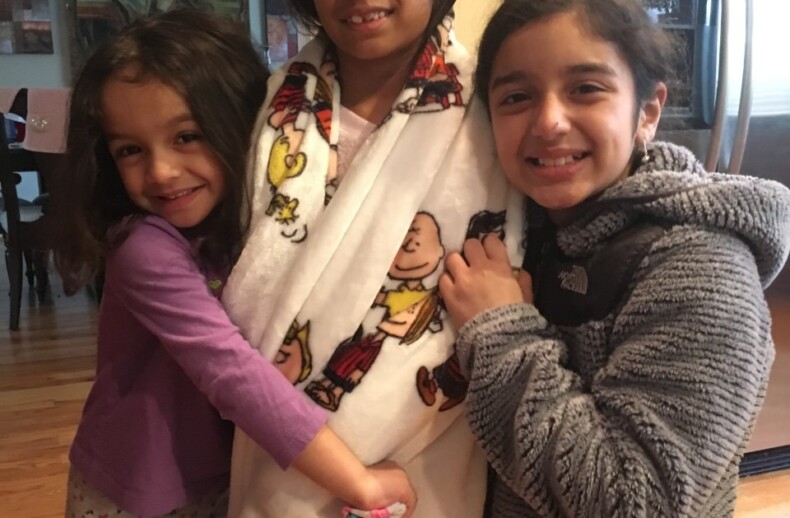 Three smiling girls stand together indoors, embodying the spirit of diversity programs. The girl in the center wears glasses and a blanket adorned with colorful cartoon characters. Her cheerful friends on either side hug her gently, illustrating the warmth fostered by SEL activities.